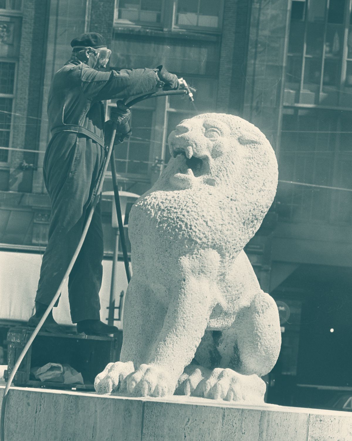 Un home amb granota de feina, dret sobre el pedestal d’una escultura de lleó de pedra, la renta amb una llança d’aigua.